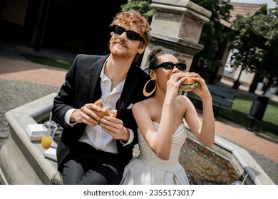 african american bride eating burger near redhead groom in sunglasses near fountain, outdoor wedding - Powered by Shutterstock