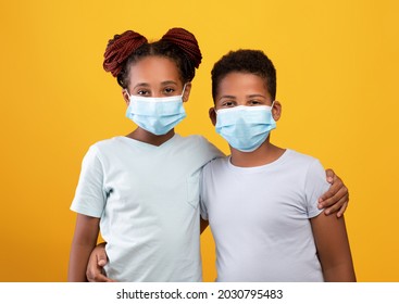 African american brother and sister wearing medical protective face masks while COVID-19 pandemic, hugging over yellow studio background. Kids and coronavirus epidemic concept