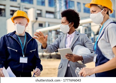 African American building contractor talking to construction engineers while inspecting project development at construction site during coronavirus epidemic. 