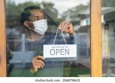 African American Business Man With Face Mask Holding The Sign For The Reopening And Looking Away Of The Place After Lockdown Quarantine. Small Business Owner Reopen And Back To New Normal Concept.