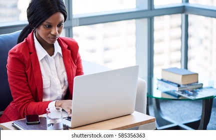 African American Business Woman Busy Looking At Her Computer Screen While Working On Her Notebook In The Lounge Of Her Business While Seated A Comfortable Couch.