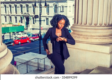 African American Business Woman Traveling, Working In New York. Holding Laptop Computer, Looking Down At Wristwatch, Young Black Lady With Braid Hairstyle Walking Into Office Building. Time Is Money. 