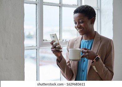 African american business woman using smartphone texting on mobile phone holding coffee standing by window