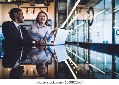 African American businessman and Asian businesswoman talking and having fun while sitting together in front of laptop at reflective counter in modern office  - Shutterstock ID 1568820307