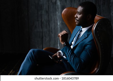 African American businessman in a blue classic suit sits in an aviator loft armchair with a phone in his hands.