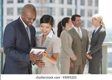 African American Businessman And Chinese Asian Businesswoman Using Tablet Computer With Interracial Group Of Business Men & Women Team.