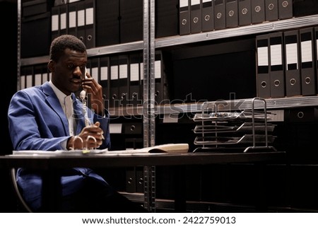 African american businessman discussing management research with remote employee using landline phone. Manager working late at night with administrative files in corporate depository