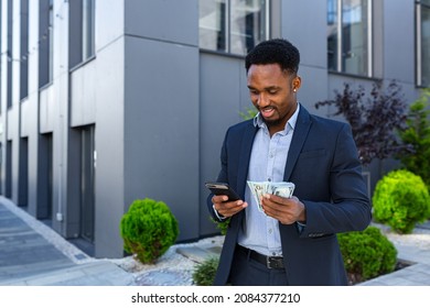 African American Businessman Standing On Street Modern Building Show Earnings Winning At Betting Cash Money With Mobile Phone In Hands. Black Man In A Suit Outdoors With Smartphone And Pack Dollars