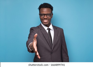 African American Businessman In Suit And Glasses Reaching Out To Shake Hands Making An Agreement With His Business Partner. Studio Shot On Blue Wall.