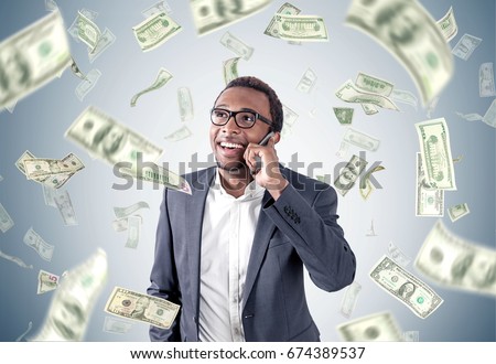 African American businessman in a suit is talking on a smartphone while standing near a gray wall with dollar bills falling around him.