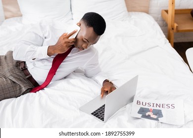 African american businessman talking on smartphone and using laptop near newspaper on bed