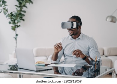 African American Businessman Uses Virtual Reality Glasses Working In Cyberspace At Laptop In Office. Modern Black Male Business Person Wearing VR Goggles Interacting With Augmented Reality.