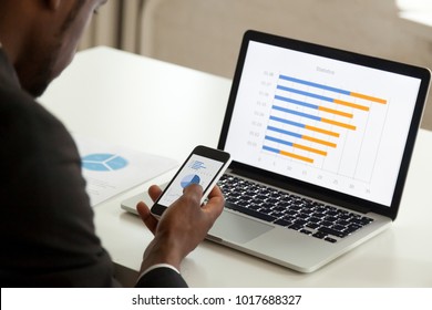African american businessman using devices for business, black office worker holding smartphone working with laptop and mobile phone, corporate statistics apps and software, over the shoulder view