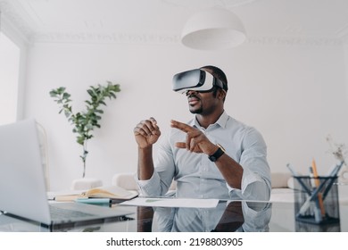 African american businessman using virtual reality glasses sitting at laptop at workplace in office. Modern black guy businessperson wearing VR goggles. Augmented Reality. Future high-tech. - Powered by Shutterstock