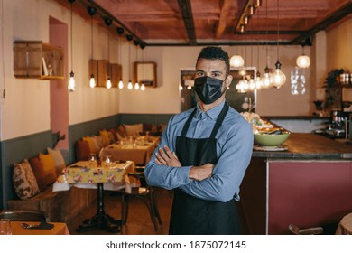 African American Businessman Wearing A Protective Black Face Mask And Standing At His Restaurant During Coronavirus. Proud Owner With His Arms Crossed And Looking At The Camera With A Defiant Gaze.