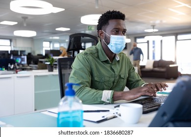 African American businessman working in modern office wearing face mask, sitting at desk using computer. Hygiene and social distancing in workplace during Coronavirus Covid 19 pandemic.
