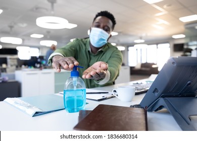 African American businessman working in modern office wearing face mask, sitting at desk using hand sanitizer. Hygiene and social distancing in workplace during Coronavirus Covid 19 pandemic. - Powered by Shutterstock