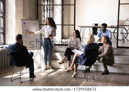 African American businesswoman coach speaker presenting statistics on flip chart at briefing, confident female mentor explaining project strategy, plan to diverse employees at corporate meeting