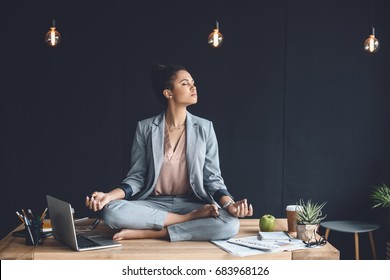african american businesswoman sitting in lotus pose on table while meditating in office