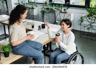 african american businesswoman sitting on desk during coffee break near colleague in wheelchair