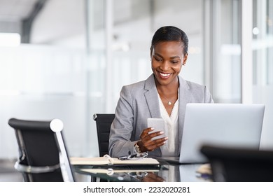 African american businesswoman using smartphone while working on laptop in office. Smiling mature black business woman checking phone while working. Successful woman entrepreneur browsing on phone. - Powered by Shutterstock
