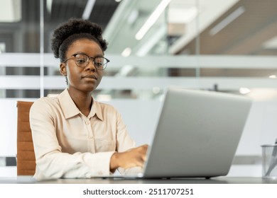 African American businesswoman wearing glasses using laptop in modern office. She appears focused and engaged with work, demonstrating professionalism and commitment in her role. Clean environment - Powered by Shutterstock