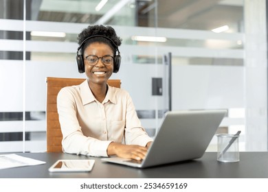 African American businesswoman wearing headphones working on laptop in modern office setting. Smiling confidently, multitasking with commitment and enjoyment. Corporate environment promotes - Powered by Shutterstock