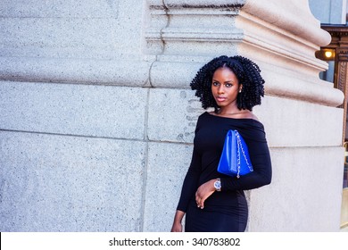 African American Businesswoman working in New York. Wearing long sleeve, slim off shoulder dress, carrying blue bag under arm, a young black lady standing on street. Filtered look with purple tint.
