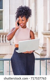 African American Businesswoman Working In New York. Young Black College Student With Afro Hairstyle Sitting On Railing In Vintage Style Office Building, Working On Laptop Computer, Making Phone Call.