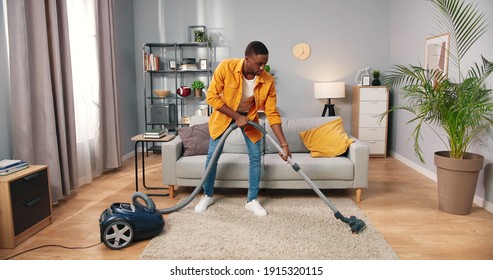 African American Busy Handsome Young Guy Vacuuming Cozy Living Room Doing Housework, Male Cleaning House Using Vacuum Keeping Home Clean, Housekeeper, Everyday Life Concept