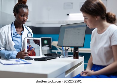 African American Cardiologist Doctor Holding Tablet Explaining Heart Xray Discussing Medical Expertise With Patient Woman During Clinical Appointment. Radiologist Working In Hospital Office.