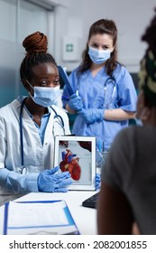 African American Cardiologist Doctor Holding Tablet Showing Heart Radiography To Sick Patient Explaining Medicaton Treatment During Clinical Appointment In Hospital Office. People With Medical Face