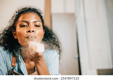 African American carpenter woman with curly hair and safety glasses blowing sawdust off wood in DIY workshop. Empowering female joiner confidently working in carpentry industry, National Carpenters - Powered by Shutterstock