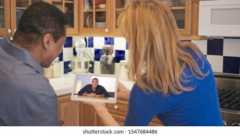 African American And Caucasian Couple In Kitchen Video Chatting With Daughter And Her Boyfriend. Older Couple Facetiming On Tablet Computer With Son And His Girlfriend