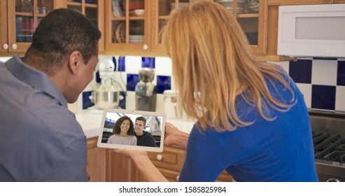 African American And Caucasian Couple In Kitchen Video Chatting With Daughter And Her Boyfriend. Older Couple Facetiming On Tablet Computer With Son And His Girlfriend