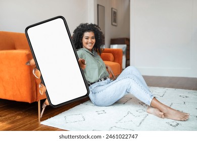 African American cheerful woman with curly hair is seated on a bright carpet floor, holding big smartphone featuring a blank screen - Powered by Shutterstock