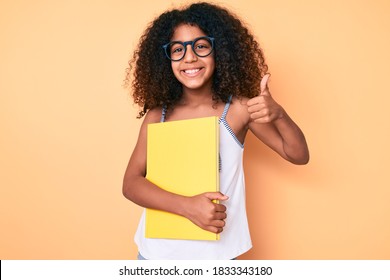 African American Child With Curly Hair Wearing Glasses And Holding Book Smiling Happy And Positive, Thumb Up Doing Excellent And Approval Sign 