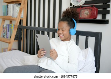 African American Child Wearing Headphones Holding Tablet Sitting On Bed. Cute Black Kid Using Smartphone Tech Listening Course Watching Video Class Elearning On Cellphone In White Bedroom At Home 