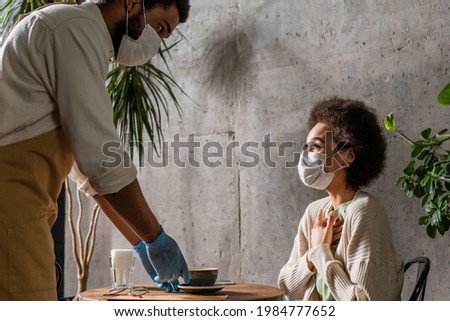 African american client in medical mask sitting near barista with coffee in cafe