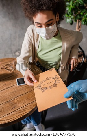 African american client in medical mask holding menu near waiter in latex glove in cafe