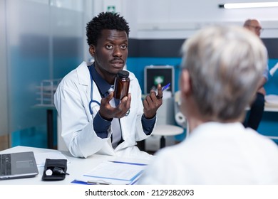 African American Clinic Expert Presenting Medicine To Elderly Patient While Explaining Side Effects. Senior Woman Talking To Young Medic About Recovery Program And Disease Stage.