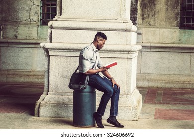 African American College Student Studying In New York. Wearing Gray Shirt, Jeans, Cloth Shoes, Carrying Shoulder Leather Bag, A Black Man Sitting On Metal Pillar On Street, Relaxing, Reading Red Book.