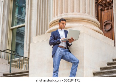 African American College Student Studying In New York, Wearing Blue Blazer, White Shirt, Gray Pants, Standing Against Column Outside Office Building, Working On Laptop Computer. 