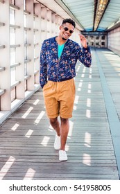 African American College Student, Wearing Blue Patterned Jacket, Yellow Brown Shorts, White Sneakers, Sunglasses, Walking On Walkway With Glass Walls On Campus In New York. Color Filtered Effect
