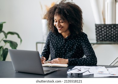 African american company manager or freelancer working in the office. Attractive confident smart curly haired woman, sitting in front of a laptop, making notes in a notebook, listens webinar, smiling - Powered by Shutterstock