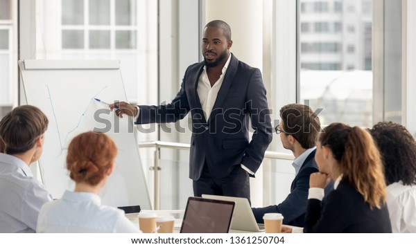 African american conference speaker coach talk to\
audience give presentation on flipchart to employees group, black\
trainer manager speaking training diverse corporate team at office\
meeting seminar