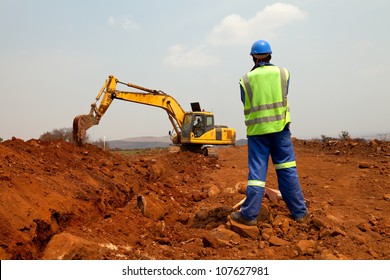 African American construction site worker wearing blue overalls and yellow high visibility safety jacket standing and watching bucket digger digging trench on sandy rocky land - Powered by Shutterstock