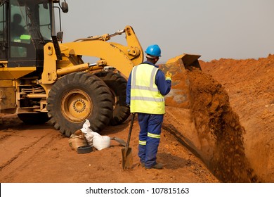 African American Construction Site Worker Wearing Blue Overalls And Yellow High Visibility Safety Jacket Standing And Watching Bucket Digger Digging Trench On Sandy Rocky Land