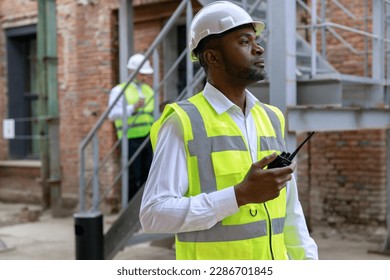 African american controller in protective hardhat using walkie talkie for regulation work and building process. Concept of safety engineer work at construction site - Powered by Shutterstock