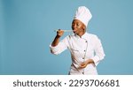 African american cook doing food taste test with spoon, sipping soup for taste testing on camera. Young gourmet chef in apron tasting restaurant meal over blue background, tasty food.